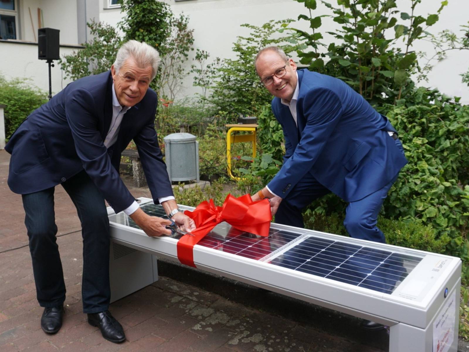 Zwei Herren in Anzug zerschneiden ein Geschenkband, dass um eine Solar-Sitzbank gewickelt ist.